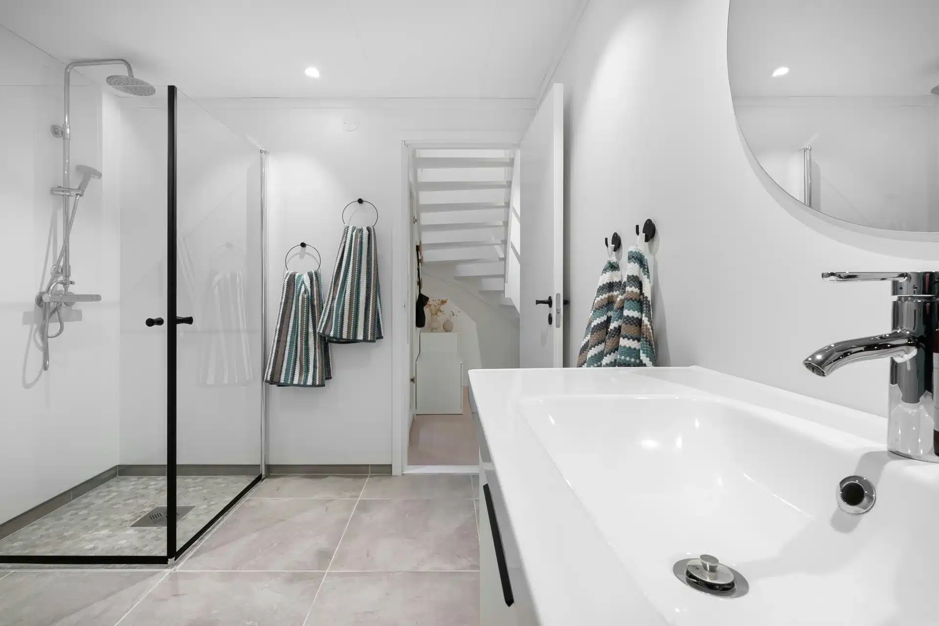 A bright, minimalist bathroom featuring a glass-enclosed walk-in shower with a rainfall head, large grey floor tiles, and a white vanity with a chrome faucet.