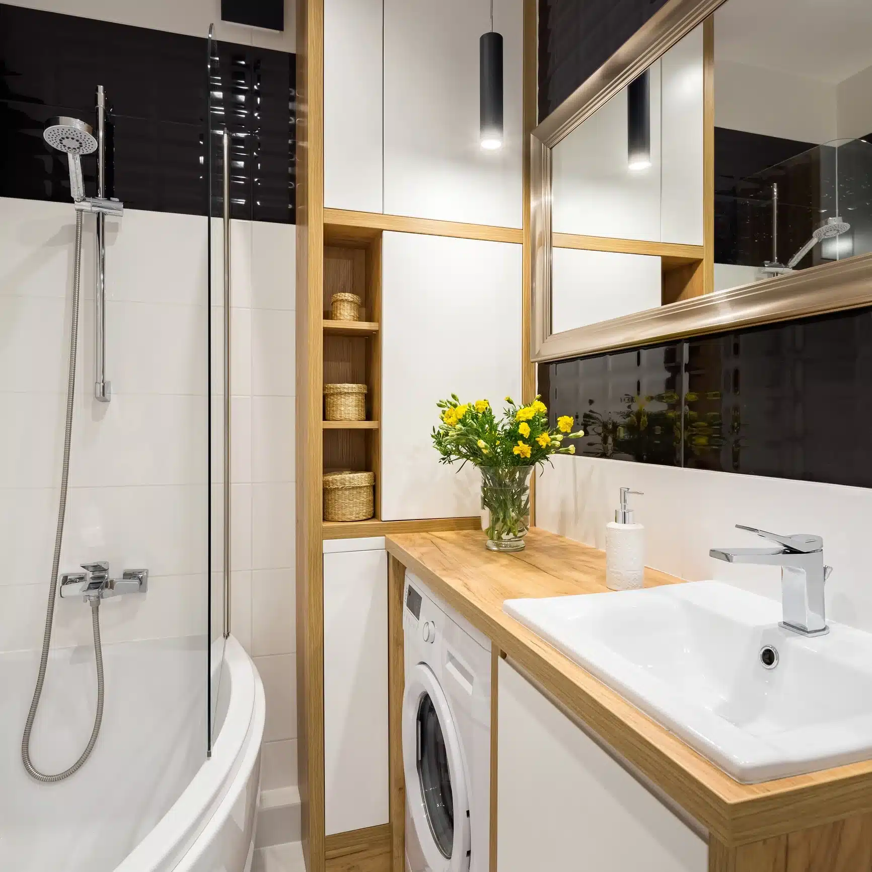 A modern, compact bathroom featuring an integrated washing machine under a wooden countertop, a white vanity sink with chrome tap, and a glass-enclosed bathtub with a chrome shower mixer.