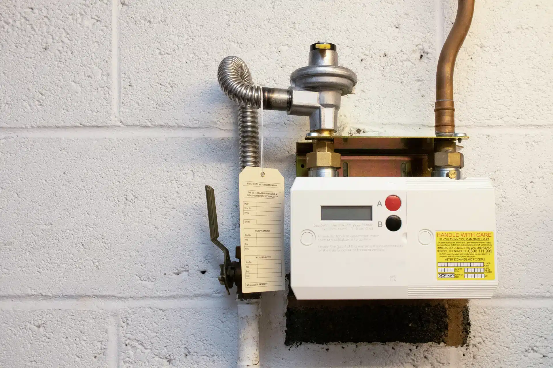 modern white smart gas meter with a digital display and emergency control valve, professionally installed against a white brick wall with copper and flexible piping.