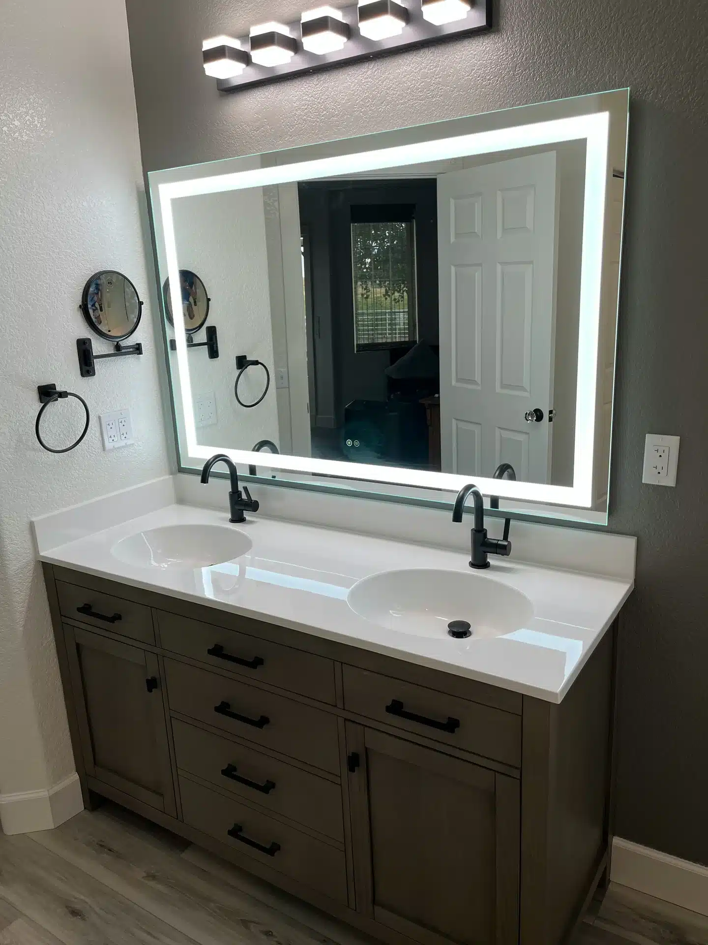 A renovated bathroom vanity featuring a double sink with white countertops, matte black gooseneck faucets, and a large rectangular LED backlit mirror