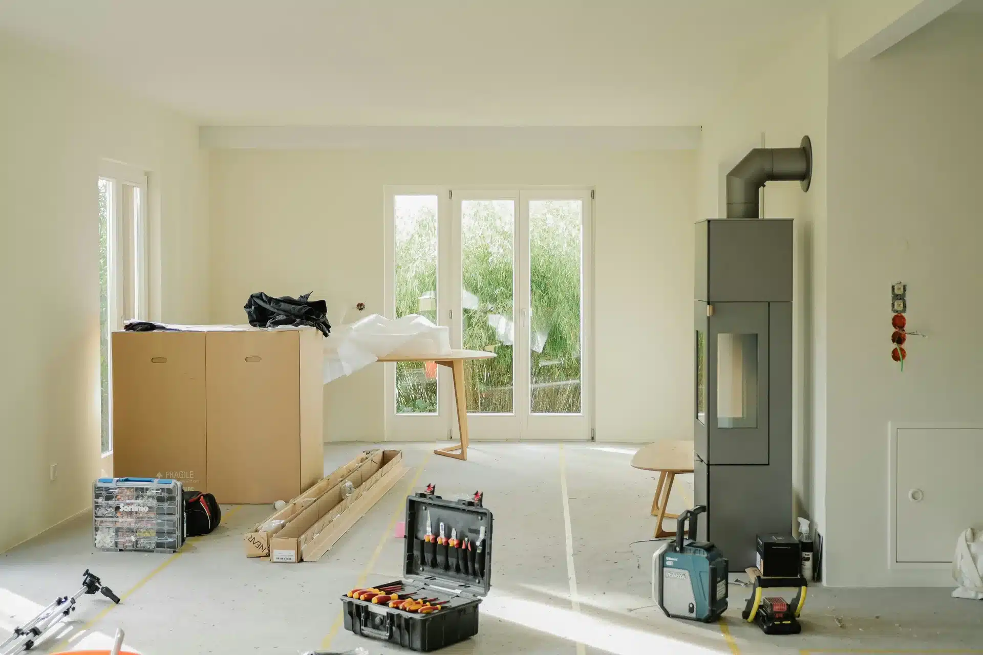 A modern grey wood-burning stove being installed in a bright, minimalist room, with professional plumbing and heating toolboxes and equipment on the floor.