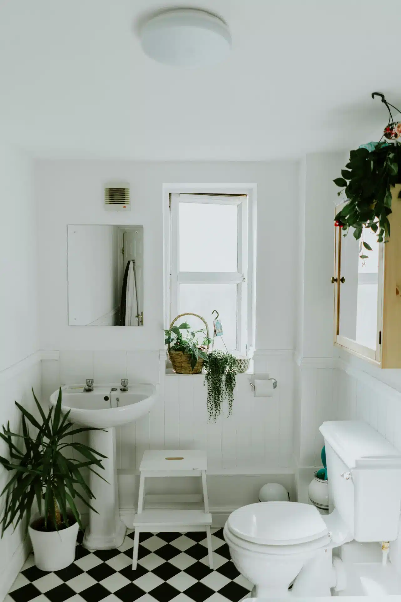 A bright, white bathroom featuring a classic pedestal washbasin with chrome taps, a matching toilet, and black and white checkered floor tiling.