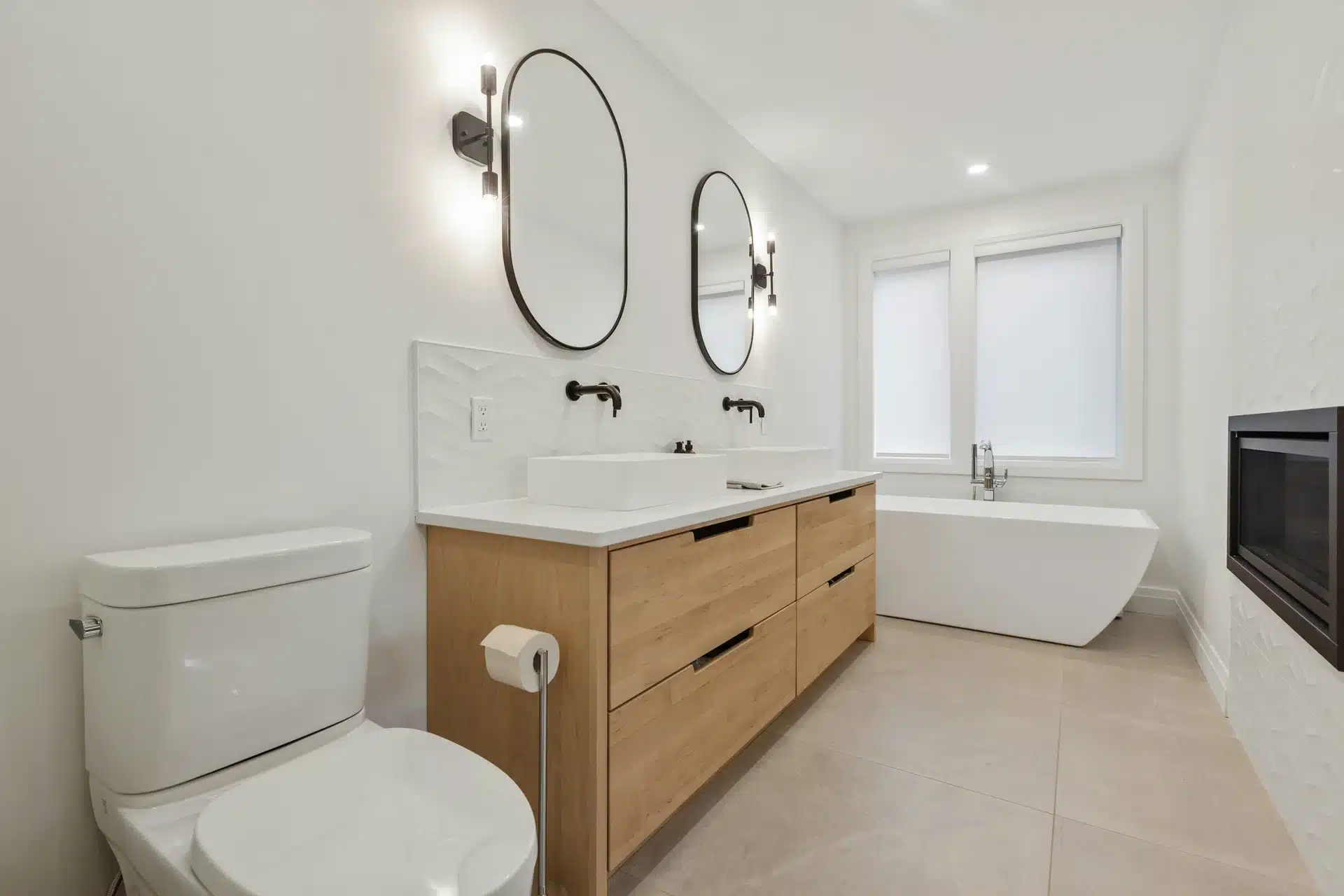 A luxury master bathroom featuring a light wood double vanity with vessel sinks, matte black wall-mounted faucets, a white toilet, and a freestanding soaking tub.