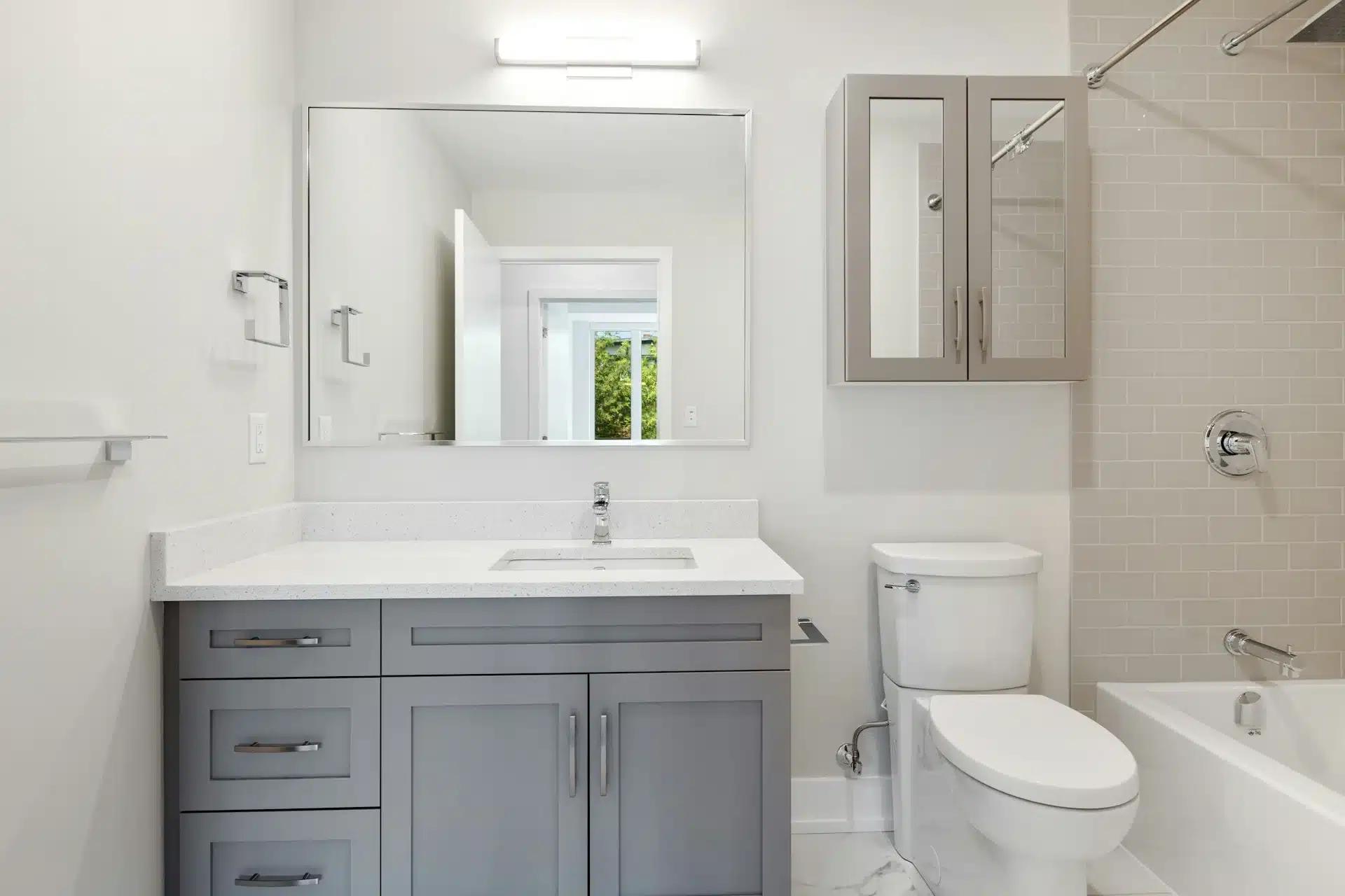 A clean, renovated bathroom featuring a grey vanity with a white countertop, a modern white toilet, and a bathtub with chrome fixtures and light tan subway tiling.