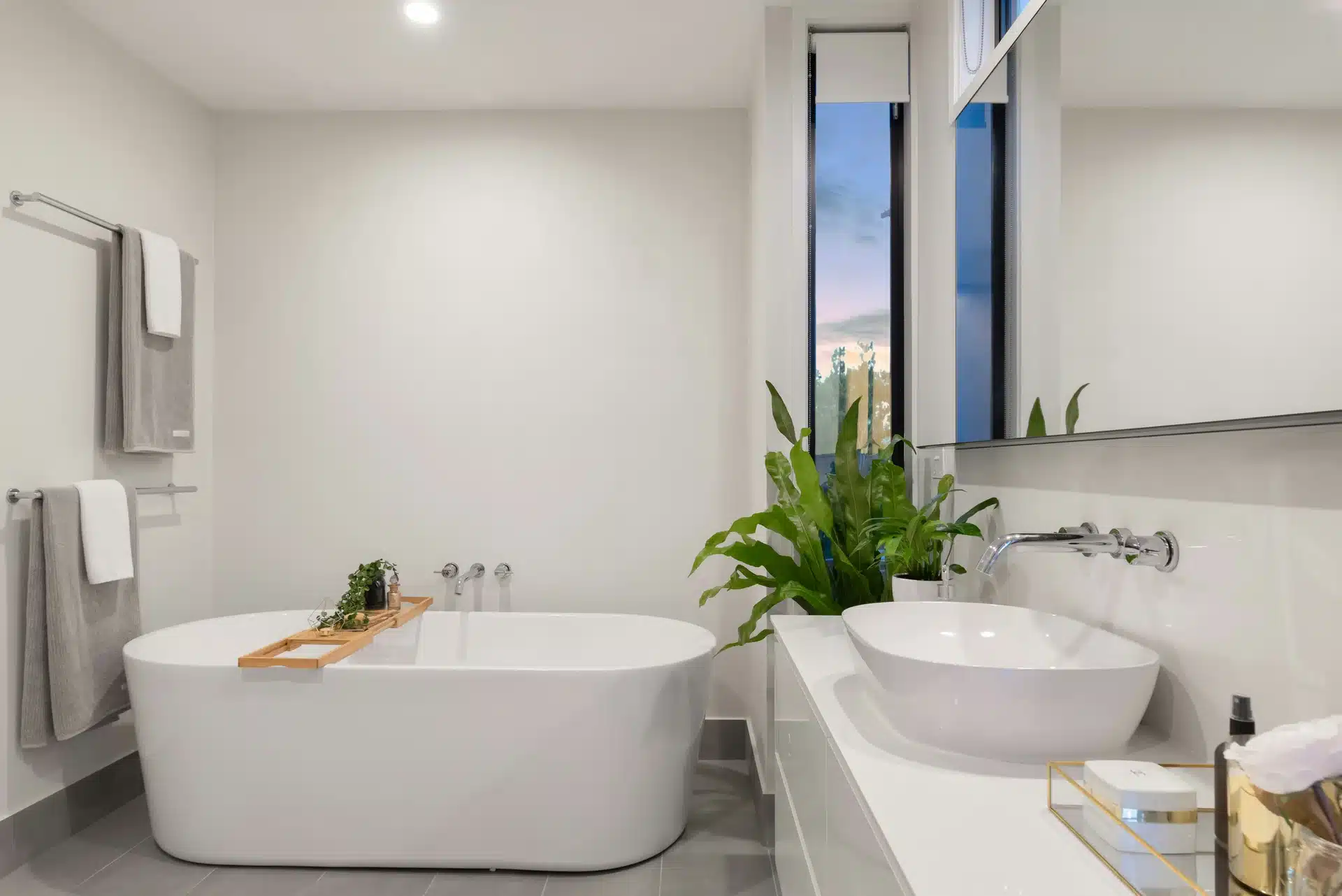 A modern white bathroom featuring a freestanding soaking bathtub with a wooden tray and a matching vessel sink with wall-mounted chrome faucets.