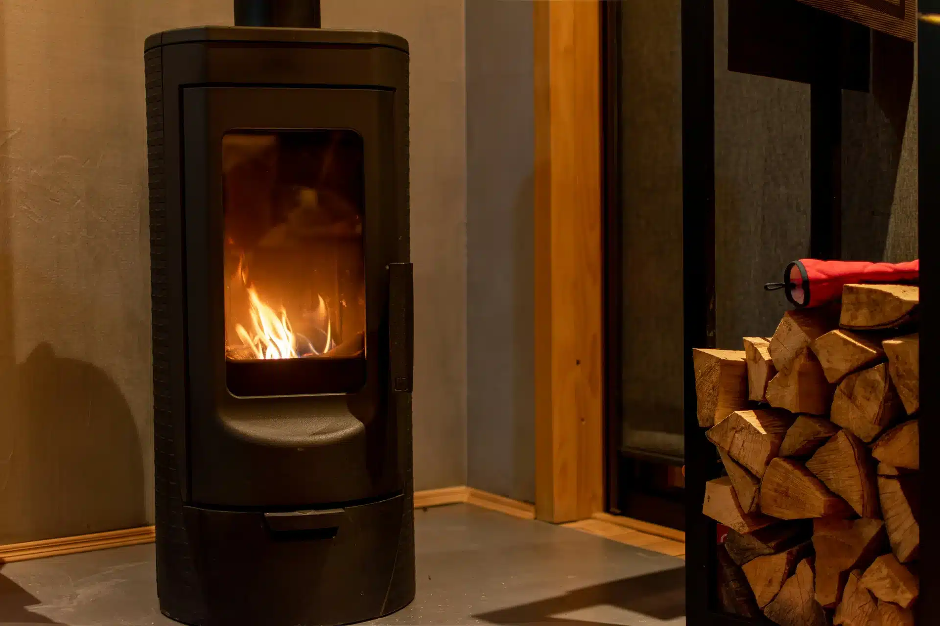 A sleek, black modern wood-burning stove with a live fire burning inside, situated in a contemporary home living area next to a stack of firewood.